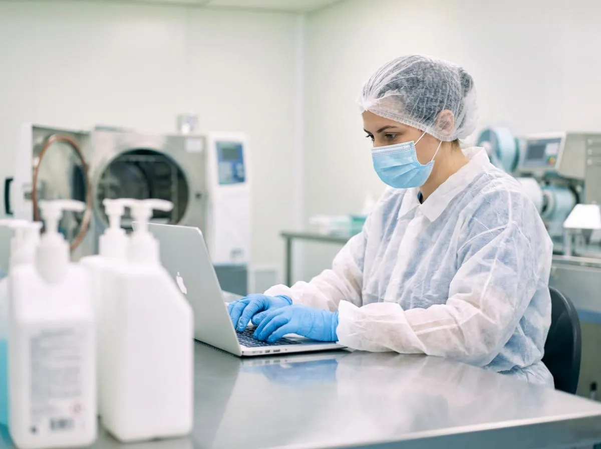 Ausbildung zum Hygienebeauftragten: Fachkraft in weißer Schutzkleidung, Mundschutz und Handschuhen arbeitet an einem Laptop in einem sterilen Laborraum neben einem Autoklaven und Desinfektionsmitteln.